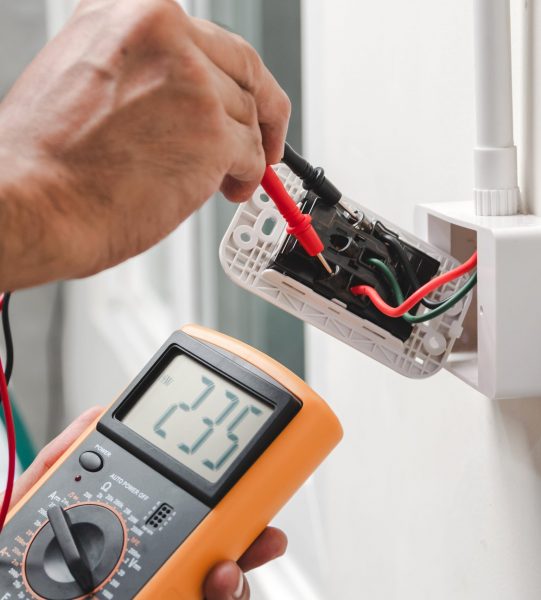 Electrician is using a digital meter to measure the voltage at the power outlet in on the wall.